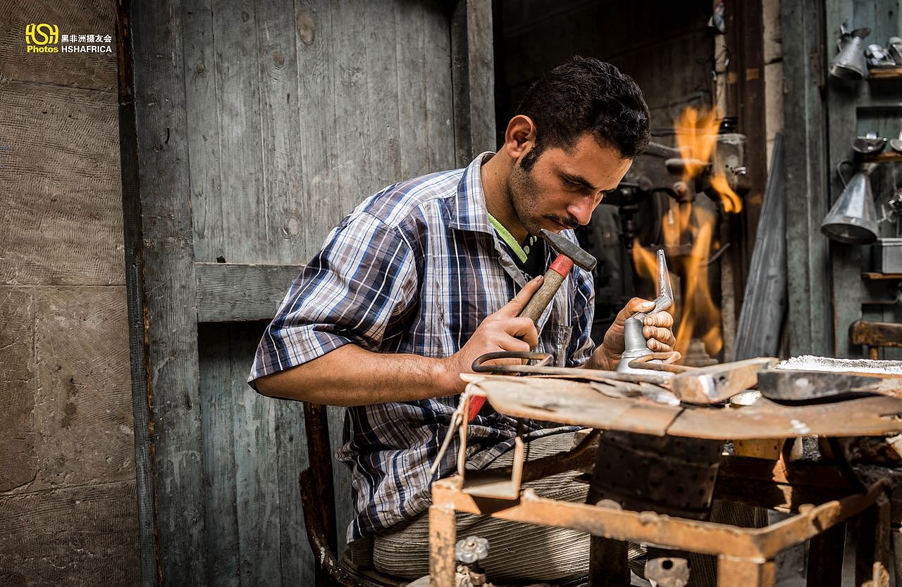 一个铁匠铺的工匠，在做铁器，旁边燃烧的煤气炉，用来加热铁锤，他用烧红的铁锤融化焊锡，给白铁皮的漏斗上锡。